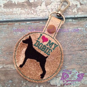 Può includere: Un portachiavi rotondo di glitter dorati con una silhouette nera di un Doberman Pinscher e il testo "Amo il mio Dober" in turchese e rosa.