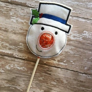 Könnte beinhalten: Ein weißer Filz-Schneemann-Lollihalter mit einem blauen Hut, grünem Stechpalmenblatt und einem roten und orangenen Lolli.