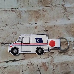 May include: Embroidered keychain featuring a white mail truck design with red and blue stripes, a blue square with a crescent moon, and a red circle. The keychain includes a metal ring for attaching keys.
