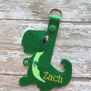 Może przedstawiać: Zielony brelok w kształcie dinozaura z metalowym kółkiem i zielonym guzikiem. Brelok jest wyszyty imieniem "Zach".
