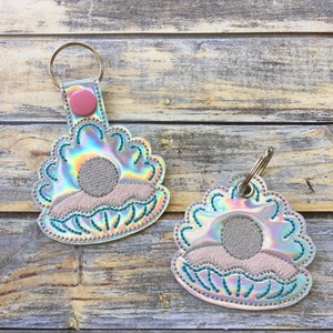 Puede incluir: Dos llaveros iridiscentes con forma de concha marina con un botón rosa en la parte superior de uno de los llaveros. Las conchas tienen un contorno azul claro y un diseño de sol blanco y rosa en el interior.