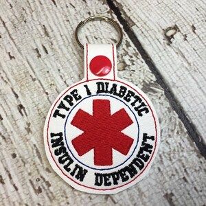 May include: White round keychain with a red medical symbol and the text "Type 1 Diabetic Insulin Dependent" in black.
