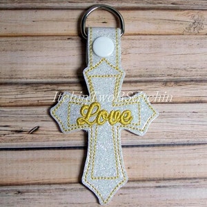 May include: A silver glitter cross keychain with a gold embroidered "Love" on it. The keychain has a silver ring and a white button.