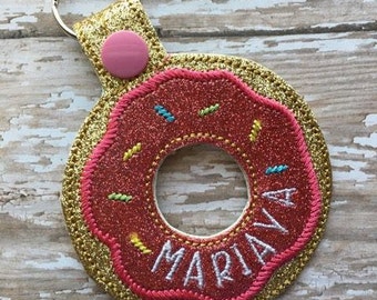 May include: A pink and gold glitter donut-shaped keychain with the name "MARIANA" embroidered in white on the donut.