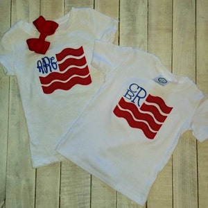 以下が含まれることがあります： 赤、白、青のアメリカ国旗のデザインが施された白いTシャツ2枚。シャツには、青い刺繍で「ARE」と「CRB」のイニシャルが旗に施されています。