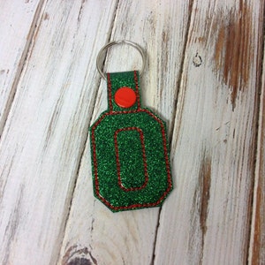 May include: Green glitter keychain with a red stitched border and a red button. The keychain is shaped like a rectangle with rounded corners.