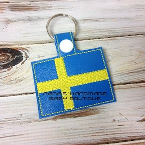 May include: A blue and yellow Swedish flag keychain with a white button and a silver key ring. The keychain is made of leather and has a stitched border. The text "Nana's Handmade Baby Boutique" is printed on the keychain.