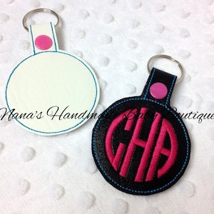May include: Two round keychains. One is white with blue stitching and the other is black with pink monogrammed letters. Both have a silver key ring, a pink button, and the text "Nana's Handmade Baby Boutique".