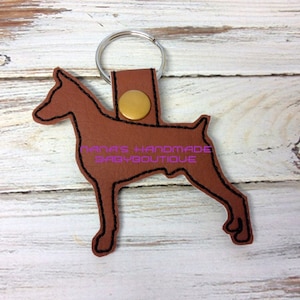Puede incluir: Llavero de cuero marrón en forma de perro Doberman Pinscher con un anillo de metal dorado y un botón dorado. El llavero tiene el texto "Nana's Handmade Baby Boutique" bordado.