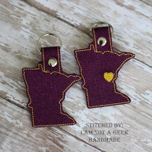 May include: Two purple glitter Minnesota state-shaped keychains with yellow stitching. The keychain on the right has a small yellow heart in the center of the state. The keychains are on a light wood background. The text "STITCHED BY: I AM NOT A GEEK HANDMADE" is below the keychains.