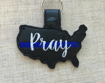 Puede incluir: Llavero de cuero negro con forma de Estados Unidos con una línea fina azul y la palabra "Pray" bordada en blanco.