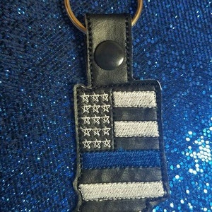 May include: A black leather keychain with a blue and white American flag design and a thin blue line. The keychain is attached to a silver key ring.