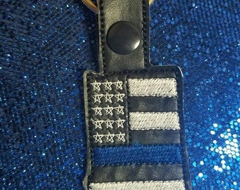 Puede incluir: Un llavero de cuero negro con un diseño de bandera estadounidense azul y blanca y una fina línea azul. El llavero está unido a un anillo de llave plateado.