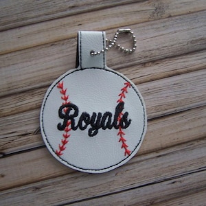 May include: White leather baseball keychain with red stitching and black embroidery that says "Royals".