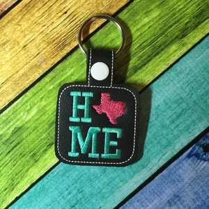 May include: A black leather keychain with a white button and a silver key ring. The keychain has an embroidered design of the word "HOME" with a pink outline of the state of Texas in the middle of the word.