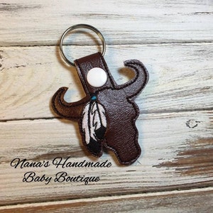 May include: Brown leather keychain with a white button and a brown buffalo skull design with a blue and white feather. The text "Nana's Handmade Baby Boutique" is visible at the bottom of the image.