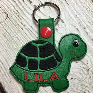 May include: Green turtle-shaped keychain with a black shell and red stitching. The keychain has a metal ring and a red button. The name "LILA" is embroidered in red on the turtle's shell.