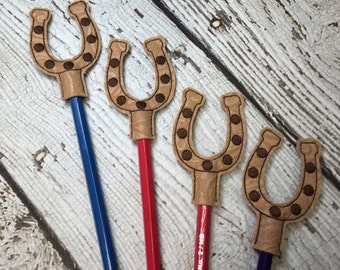 May include: Four brown felt pencil toppers shaped like horseshoes with brown polka dots. The toppers are on pencils of different colors: blue, red, purple, and a natural wood pencil.