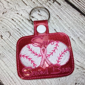 Peut inclure: Un porte-clés sur le thème du baseball rose et blanc avec le texte "Save Second Base". Le porte-clés présente deux balles de baseball blanches avec des coutures roses et un ruban rose au centre.