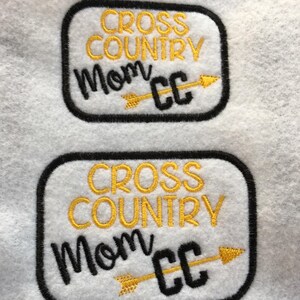 Puede incluir: Dos parches blancos con bordes negros y bordados dorados. El parche superior dice "Cross Country Mom CC" con una flecha apuntando hacia la derecha. El parche inferior dice "Cross Country Mom CC" con una flecha apuntando hacia la derecha.