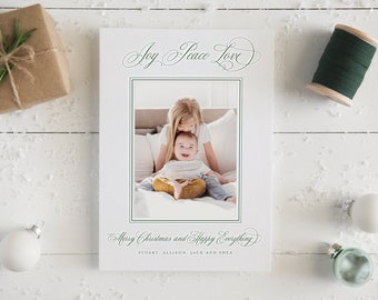 Op de afbeelding: Een witte kerstkaart met een familiefoto, omringd door groene tekst met de tekst "Joy Peace Love" en "Merry Christmas and Happy Everything". De kaart is versierd met kerstballen, een ingepakt cadeau en een spoel groen garen.