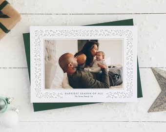 May include: A holiday card with a family photo inside a floral border. The card is on a green envelope, with a wrapped gift and ornaments nearby. The text reads "Happiest Season of All".