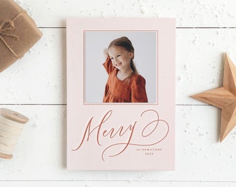 Puede incluir: Una tarjeta navideña rosa con una foto de una niña sonriente con un vestido naranja. La tarjeta presenta la palabra "Merry" en una escritura elegante, junto con el apellido y el año. También se ve un regalo envuelto y una estrella de madera.
