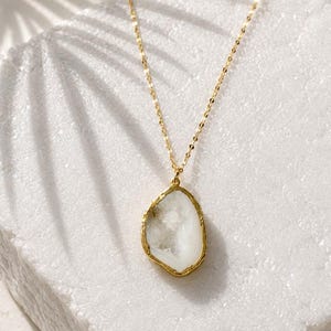 May include: A gold chain necklace featuring a pendant with an irregular, organic-shaped white stone set in a gold-toned metal frame. The pendant has a textured, crystalline center and a smooth, polished edge, suspended from a delicate gold link chain.