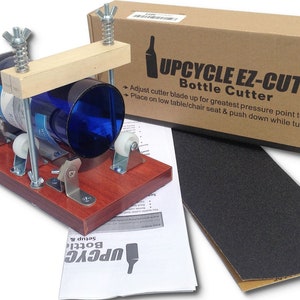 Op de afbeelding: Een houten flessensnijder met een blauwe glazen fles erin. De snijder heeft twee rollen en een metalen mes. De doos voor de snijder is op de achtergrond. De tekst op de doos luidt "Upcycle Ez-Cut Bottle Cutter".