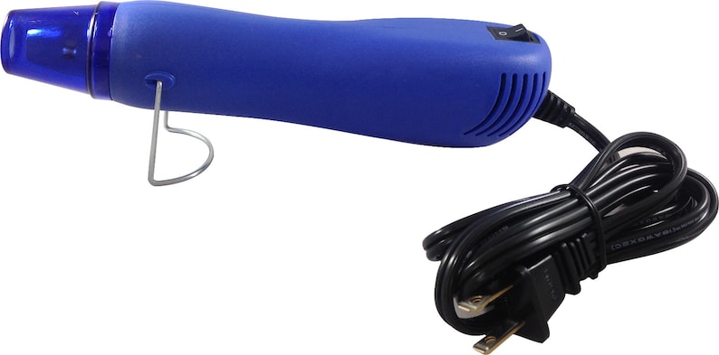 Puede incluir: Una pistola de calor azul con un cable de alimentaci&oacute;n negro y un enchufe de dos clavijas. La pistola de calor tiene una boquilla transparente y un soporte de metal plateado. El interruptor de encendido/apagado est&aacute; situado en la parte superior de la herramienta.