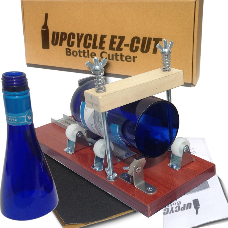 Op de afbeelding: Een houten flessensnijder met een blauwe glazen fles erin. De snijder heeft twee metalen rollen en een houten handvat. De doos voor de snijder is op de achtergrond. De tekst op de doos luidt "Upcycle Ez-Cut Bottle Cutter".