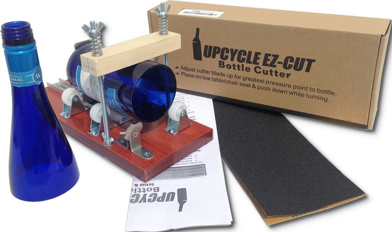 Op de afbeelding: Een houten flessensnijder met een blauwe glazen fles erin. De snijder heeft twee rollen en een metalen mes. De doos voor de snijder is op de achtergrond. De tekst op de doos luidt "Upcycle Ez-Cut Bottle Cutter".