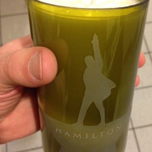 Puede incluir: Vela verde oliva en un vaso reciclado. El vaso presenta una silueta esmerilada de una persona con el brazo levantado y la palabra "HAMILTON" debajo. La vela tiene cera blanca y una sola mecha.