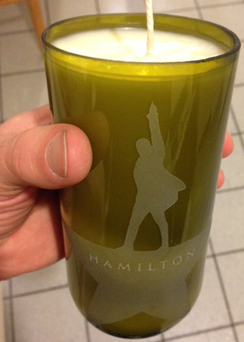 Puede incluir: Vela verde oliva en un vaso reciclado. El vaso presenta una silueta esmerilada de una persona con el brazo levantado y la palabra "HAMILTON" debajo. La vela tiene cera blanca y una sola mecha.
