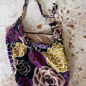 May include: A black floral shoulder bag with a cream-colored interior. The bag features large, stylized flowers in shades of purple, olive green, and tan. The strap is made of the same floral fabric and is attached to a silver metal ring.