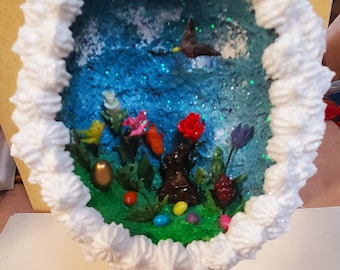 May include: A white ceramic Easter egg with a whimsical scene inside. The scene features a blue sky with clouds, a flying bird, a brown bunny, flowers, and green grass. The egg is topped with pink flowers and sits on a white pedestal with pink trim.