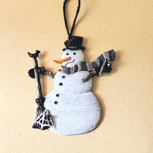 May include: A white snowman ornament with a black top hat, a black scarf, and a broom with a spiderweb. The snowman is holding the broom in its right hand and has a carrot nose.