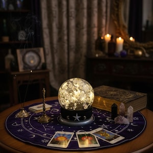 May include: A lit crystal ball sits on a table with tarot cards, crystals, and a large book. The table is covered with a purple cloth with celestial designs. Incense burns nearby, creating a mystical atmosphere.