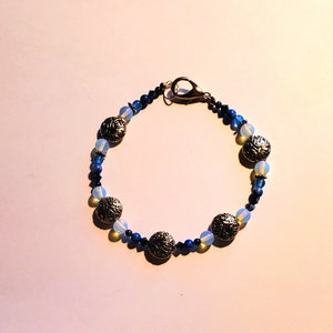 May include: A blue and silver beaded bracelet with a clasp closure. The bracelet features round, silver beads with a decorative design and blue glass beads.