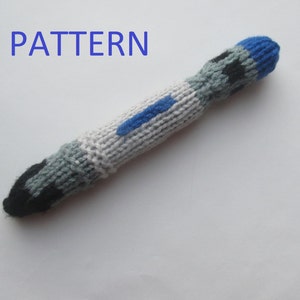 May include: A knitted toy in a cylindrical shape, featuring a colour scheme of grey, black, and blue. It has a blue tip and a blue stripe. The word "PATTERN" is written in blue at the top of the image.
