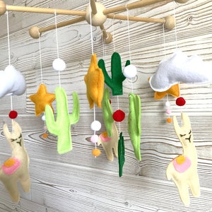 May include: A baby mobile with a desert theme. It features two white clouds, two yellow stars, two green cacti, and two cream-colored llamas with pink accents. The mobile is suspended from a wooden frame.