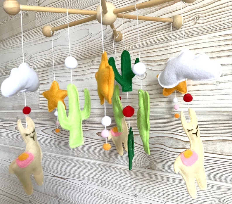 May include: A baby mobile with a desert theme. It features two white clouds, two yellow stars, two green cacti, and two cream-colored llamas with pink accents. The mobile is suspended from a wooden frame.