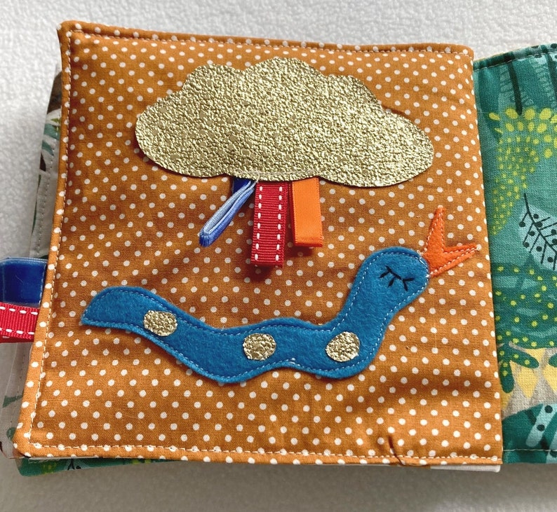 May include: A brown fabric book with a blue and orange worm, a gold glitter cloud with red and orange ribbons, and a green fabric background with a leaf pattern.
