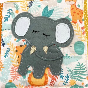 May include: A gray felt elephant with closed eyes and a gold glitter trunk. The elephant is on a white fabric background with a jungle animal print.