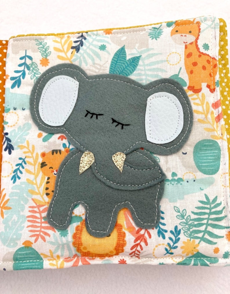 May include: A gray felt elephant with closed eyes and a gold glitter trunk. The elephant is on a white fabric background with a jungle animal print.