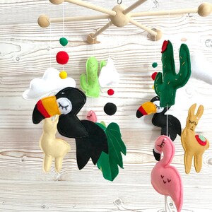 May include: A wooden baby mobile with a toucan, llama, flamingo, cactus, and palm leaf hanging from it. The mobile is painted in bright colors, including green, yellow, red, pink, and black.