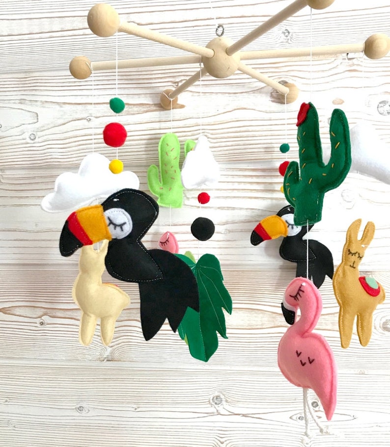 May include: A wooden baby mobile with a toucan, llama, flamingo, cactus, and palm leaf hanging from it. The mobile is painted in bright colors, including green, yellow, red, pink, and black.