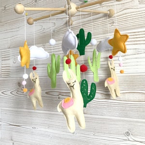 May include: A baby mobile with a white, green, and yellow color scheme. The mobile features two white llamas, green cacti, white clouds, and yellow stars. The mobile is suspended from a wooden frame.