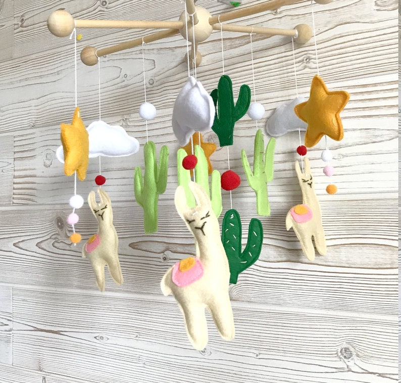 May include: A baby mobile with a white, green, and yellow color scheme. The mobile features two white llamas, green cacti, white clouds, and yellow stars. The mobile is suspended from a wooden frame.
