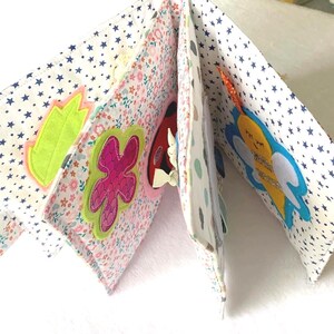 May include: A fabric baby book with appliqu&eacute;d designs. The pages display floral and star patterns, with felt shapes of a leaf, a flower, a ladybug, and a bee. The book is designed to stimulate a baby's senses with its textures and bright colours.